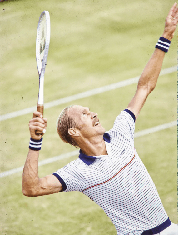 Stan Smith mid-serve during a 1970s tennis match, wearing striped athletic apparel and wristbands, with a wooden racket raised—an archival image representing his consultation with Dr. Mike Marshall on biomechanics and pitching reform principles.